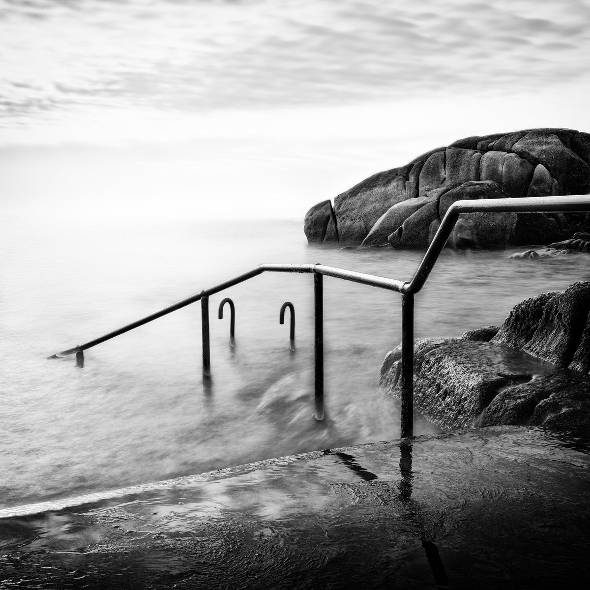 The Crossing — two iron handrails with curved ends descending into silky long-exposure coastal water with granite rocks, black and white fine art photography by Roy Fraser