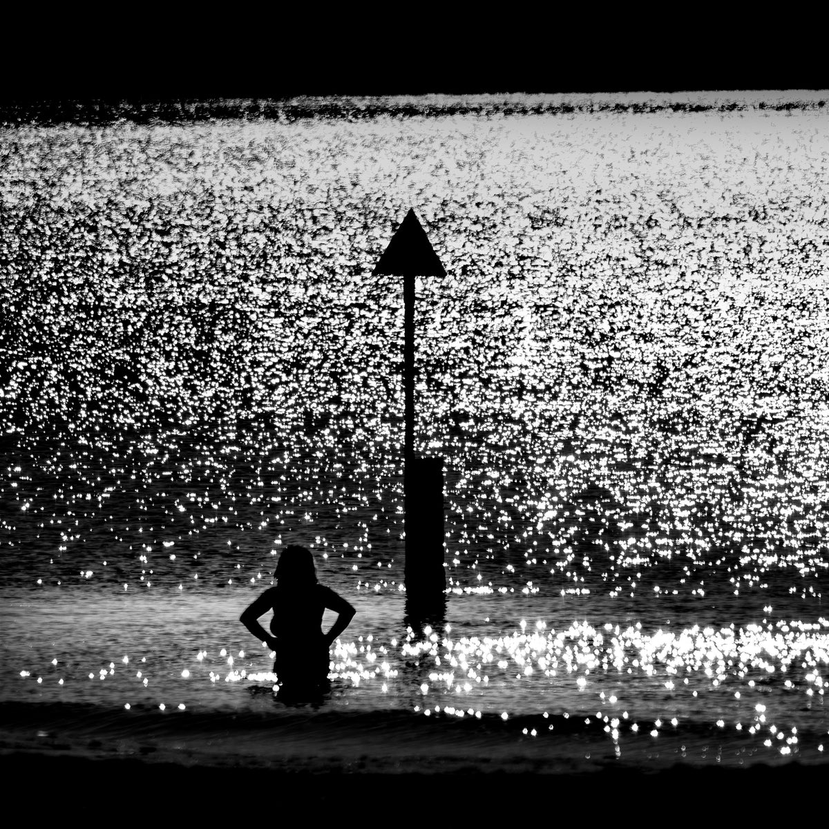 Chinmoku no Kishi — woman waist-deep in sparkling sea beside triangular beach marker, black and white fine art photography