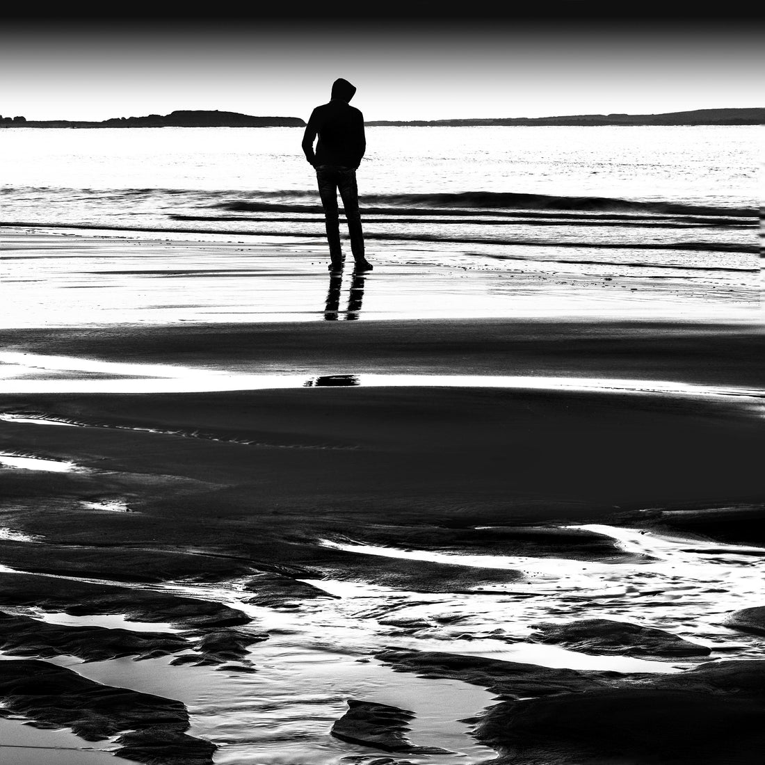 A solitary person standing still on a quiet shoreline in a minimalist landscape.