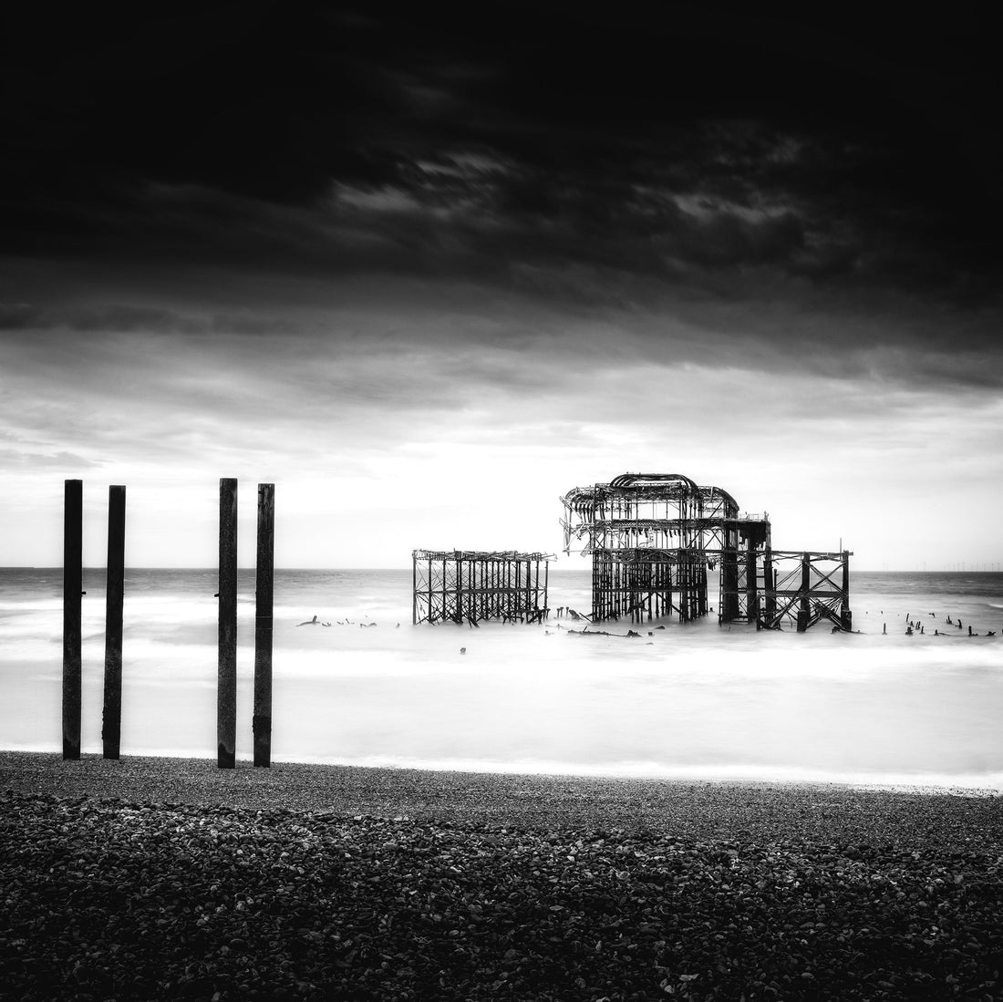 Black-and-white shoreline photograph with dark posts beside the skeletal remains of a pier.