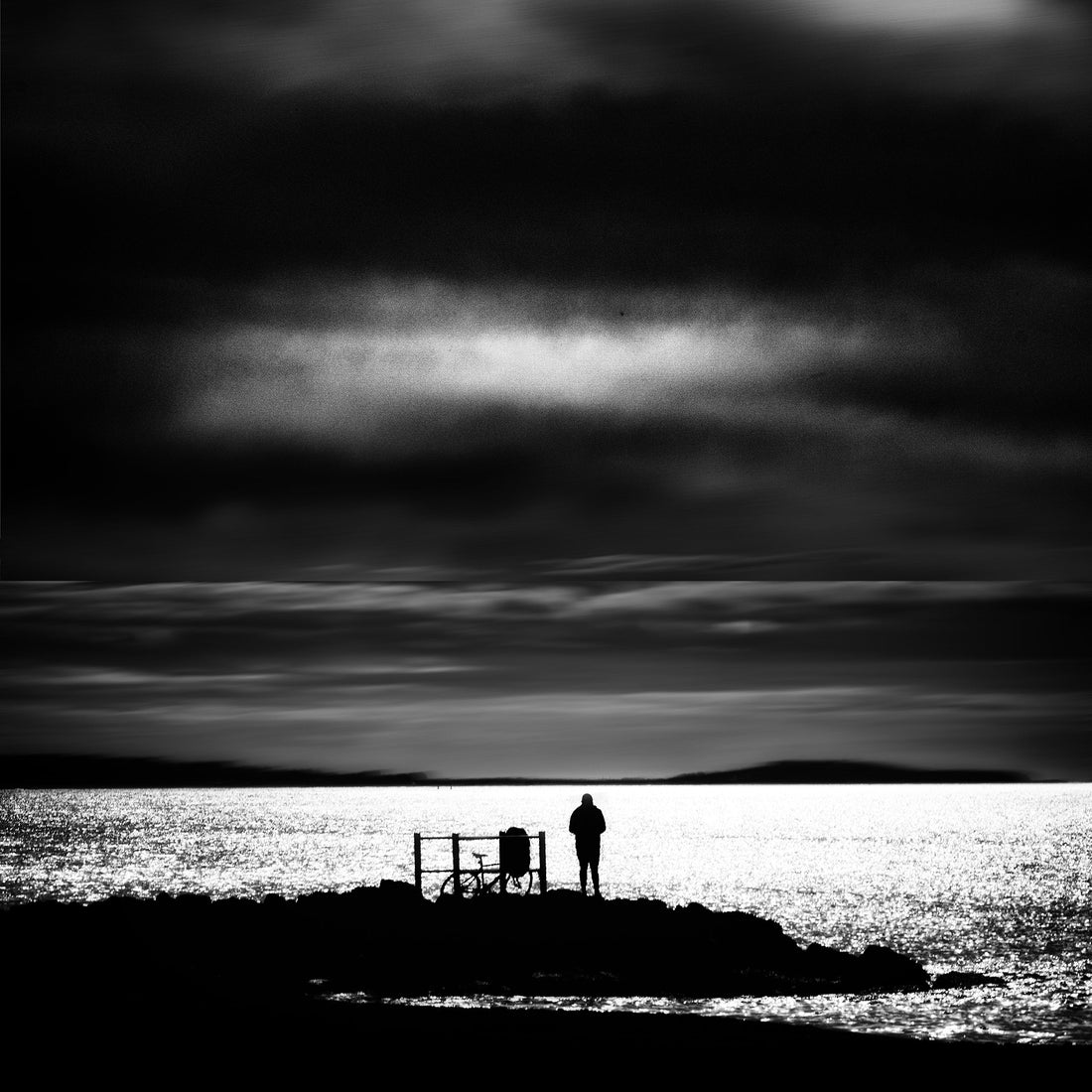 Lone figure standing beside a bicycle on a rocky coastal outcrop, silhouetted against a dramatic stormy sky — Dorset coast