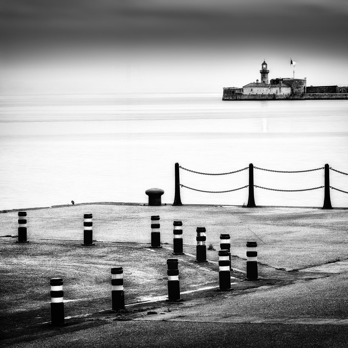 The Watch — harbour quayside with striped bollards and chain-linked posts, lighthouse and fortified building on the horizon across a silky long-exposure sea, black and white fine art photography by Roy Fraser